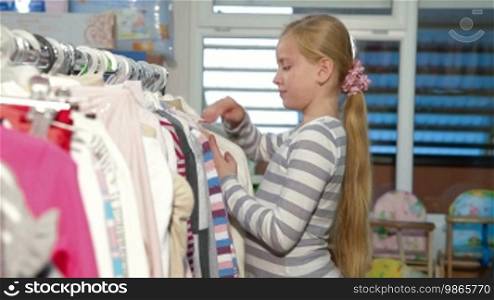 Little blond girl shopping for clothes in a clothing store, looking at a sweater. Side view