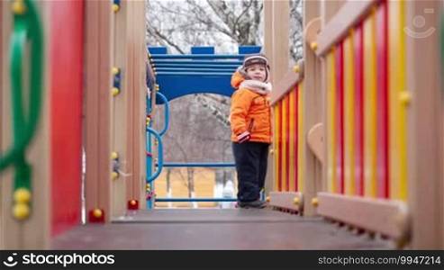 Little boy in warm clothes running toward the camera on playground equipment outdoors. Outdoor activity for children