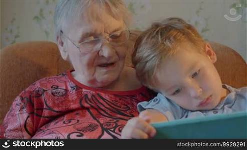 Little boy is sitting on his grandmother's lap and showing her something on a tablet PC.