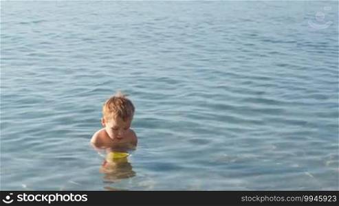 Little boy playing in the shallow sea standing waist high in the water with his plastic toys bucket and shovel in the summer sun, with copyspace