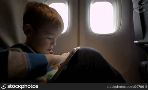 Little boy playing on a tablet computer sitting by the window in a plane