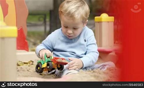 Little boy playing with toy tractor in the playground. He is taking sand with his toy and then emptying it.