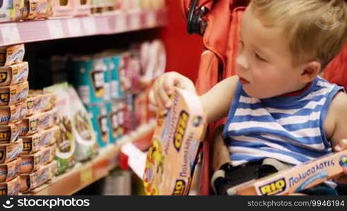 Little boy sitting in the baby carriage and taking tasty things from shelf in the shop. He taking more and more.