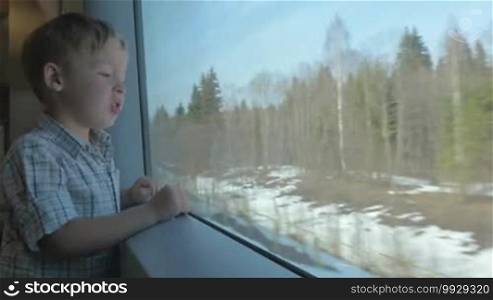 Little boy standing by the big window in moving train. He looking at passing winter or early spring landscape with bare trees, green fir-trees and snow