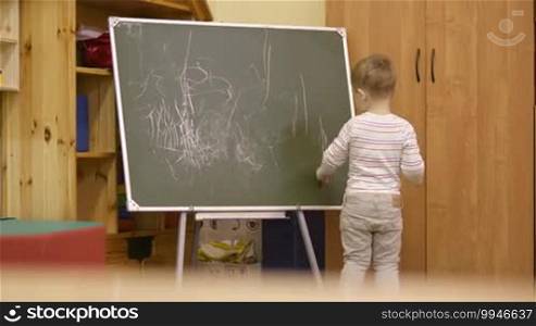 Little boy standing with his back to the camera drawing on a chalkboard at kindergarten during class as he expresses his creativity