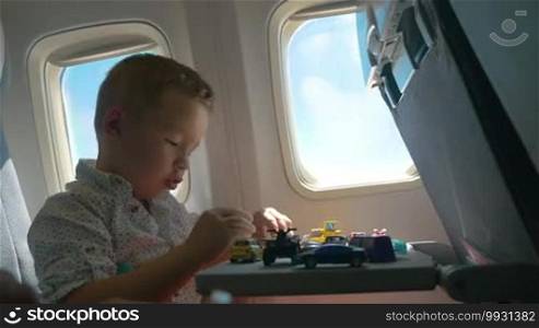 Little boy traveling by air. He is sitting by the window and playing with a toy plane, with many cars on the table in front of him. Entertaining during the flight