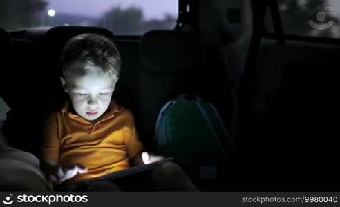 Little boy traveling on backseat of a car at night and using touch pad to entertain himself during the trip