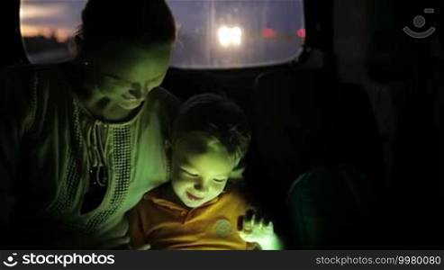 Little boy traveling with his mother on the backseat of a car at night and using a touchpad for entertainment