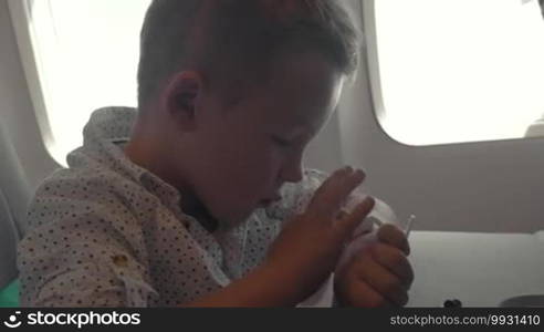 Little boy using touchscreen smart watch in the plane, he sitting by the illuminator leaning on the table. Modern generation and new technologies