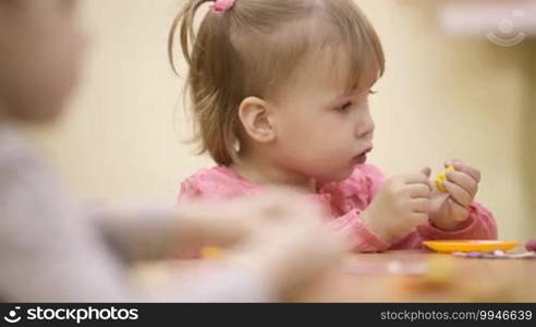 Little girl sitting at the table in the nursery and playing with plasticine. Other children sitting next to her.