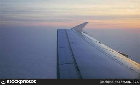 Looking through the illuminator of a flying plane at sunset. View of the wing, sky, and white clouds below