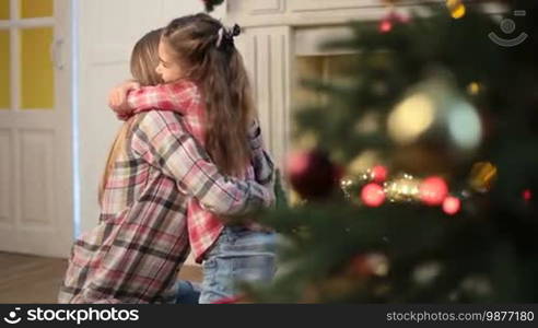 Lovely daughter with long brown hair embracing and caressing her affectionate mother while family relaxing on Christmas eve at home. Little cute girl expressing her love and affection to mom during winter holidays over xmas decorated room background.