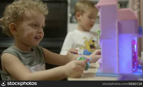 Lovely smiling little boy playing with toy plastic house, another child playing near him. Happy childhood