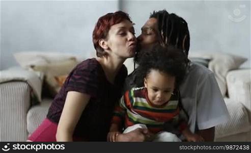 Loving affectionate mixed couple's sensual kiss while sitting on the floor with curly son in modern apartment. Happy interracial family with mixed race child spending leisure at home. Caucasian mother kissing African American father with dreadlocks.