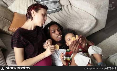 Loving diverse family with mixed-race toddler child eating cookies in the living room. Top view. Cheerful African American father with dreadlocks and Caucasian pregnant mother feeding cookies to their little boy and laughing. Slow motion.