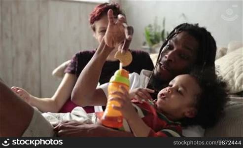 Loving interracial parents trying to convince their curly mixed-race toddler son to drink juice from a feeding bottle at home. Affectionate African American father with dreadlocks fooling around, trying to give a drink to his little kid. Slow motion.