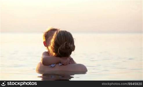 Loving mother playing with her small son in the sea holding him out of the water, hugging and kissing him backlit by the setting sun