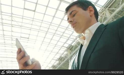 Low-angle shot of a young businessman using a smartphone in a modern building with a glass roof. He's brightly lit with sunlight.