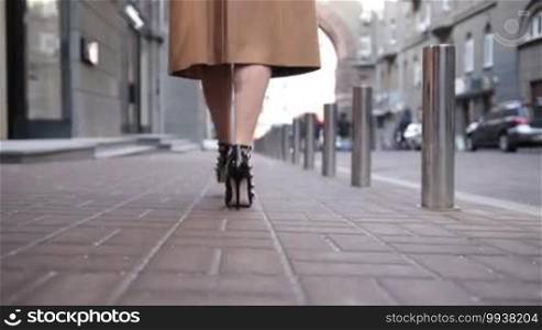 Low angle view of a business woman in black high heel shoes walking away in the city street close-up. Rear view of female beautiful legs walking on the sidewalk. Slow motion.
