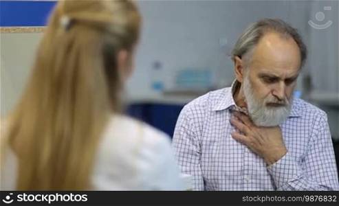 Male senior patient with beard visiting a doctor at the medical office. Elderly ill patient coughing and explaining his symptoms to female physician during checkup in doctor's office.