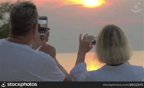 Man and woman are standing on the beach and shooting sunset at their smartphones. They are turned backs to the camera.
