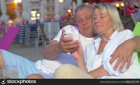 Man and woman hugging on beach while looking at phone, smiling and talking.