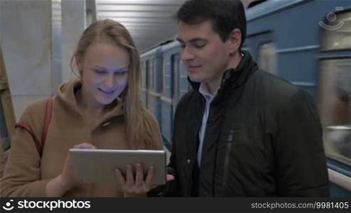Man and woman with touchpad in the underground. Woman showing something on the pad and they are discussing it. Leaving train in the background