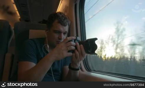 Man having a train journey. He is listening to music and looking out the window after taking a video of outside scenes at sunset