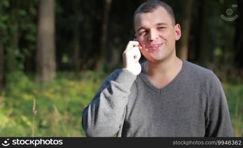 Man laughing as he chats on his mobile phone while standing outdoors in a lush green park with copyspace