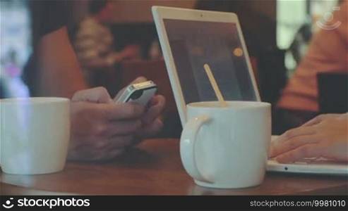 Man uses smartphone, woman works with laptop while drinking coffee in cafe, faces are invisible
