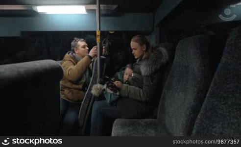 Man with cell taking video of woman with son playing on smartphone while traveling by bus in the evening