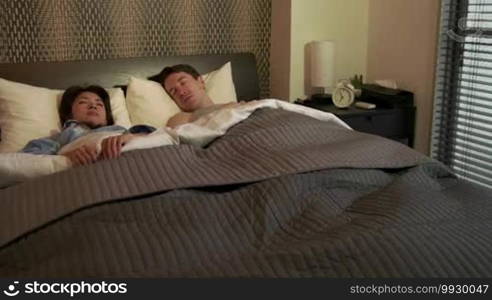 Married people, marriage relationship, husband and Asian wife at home. Early morning with tired man snoozing alarm clock, waking up and Japanese woman staying in bed. Interracial couple lifestyle