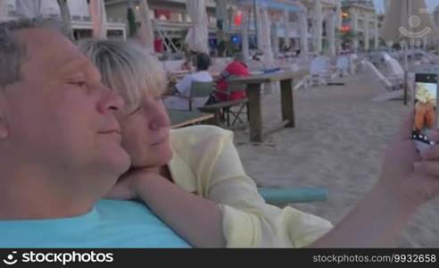 Mature couple is cuddling in beach lounger and taking selfie shot at smartphone.