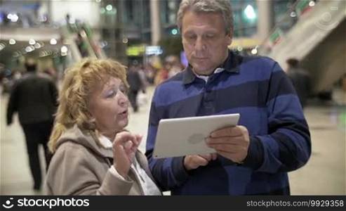 Mature couple is standing in some public place, man is holding tablet PC in hands, woman is explaining something emotionally.