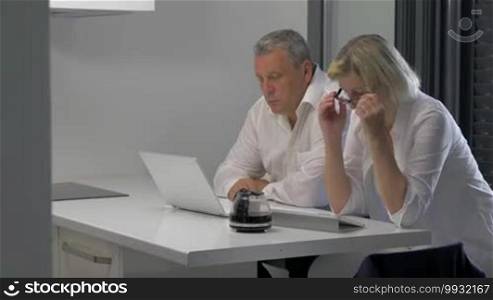 Mature man and woman with laptop and tablet PC working on business indoors. They are drinking coffee while doing their job