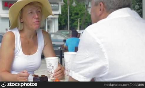 Mature woman and man are sitting in street coffeehouse, drinking coffee from paper cups and eating cakes.