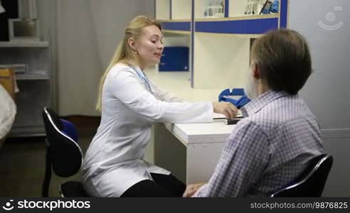 Medical exam. Confident smiling female doctor in lab coat checking senior man's temperature with electronic thermometer in doctor's office during checkup. Sick old man measuring body temperature during visit a hospital. Slow motion.