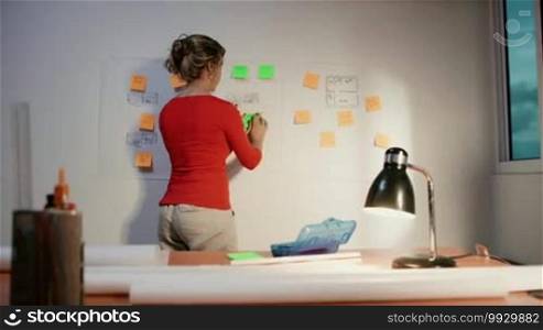 Medium shot of a blond woman working as an architect, sticking an adhesive note to a blueprint. She sits back and contemplates the missing tasks to complete the housing project. Dolly shot