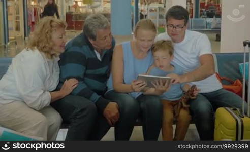 Members of a big family are watching photos from the vacation on a tablet PC while sitting in the waiting room of an airport or railway station.