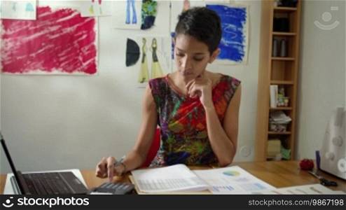 Money and financial planning, young Hispanic self-employed woman checking bills and doing budget with calculator, computer and papers in fashion design studio. Sequence