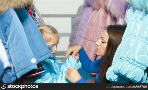 Mother and daughter shopping for winter clothes in a clothing store, child trying on a down-padded coat
