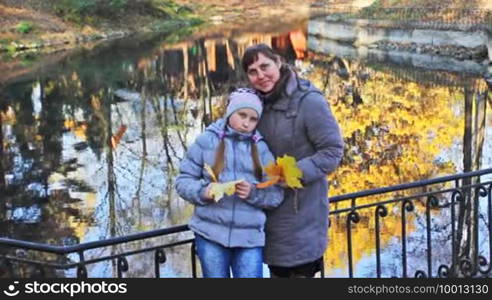 Mother and daughter with yellow leaves in their hands stand hugging near lake at beautiful autumn city park, closeup