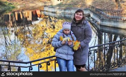 Mother and daughter with yellow leaves in their hands stand hugging near lake at beautiful autumn city park