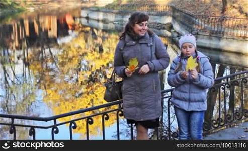 Mother and her daughter with yellow leaves in their hands stand near lake at beautiful autumn city park