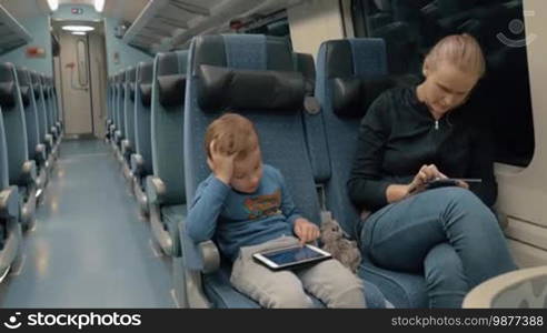 Mother and kid having night express train ride. Woman spending time with smartphone and charging it while boy playing on digital tablet