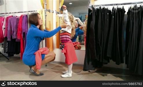Mother and little daughter shopping for girls clothes in a clothing store, trying on sweater and raincoat