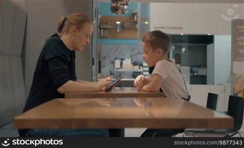 Mother and son sitting at the table in a cafe and using a mobile. Child asking mom to find something for him on the internet