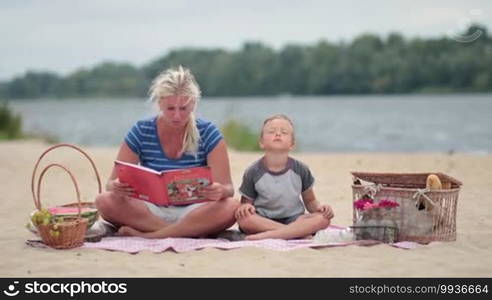 Mother and son sitting on a colorful blanket and reading a book during a picnic on the beach. Family spending time together outdoors.