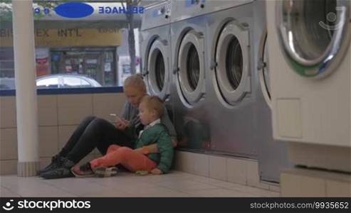 Mother and son waiting in the laundry. They are sitting on the floor near the washing machines and using a smartphone