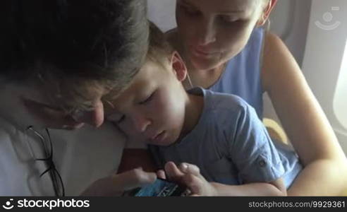 Mother, father, and little son in airplane. Father unblocking touchscreen smartphone and giving it to the son to listen to music